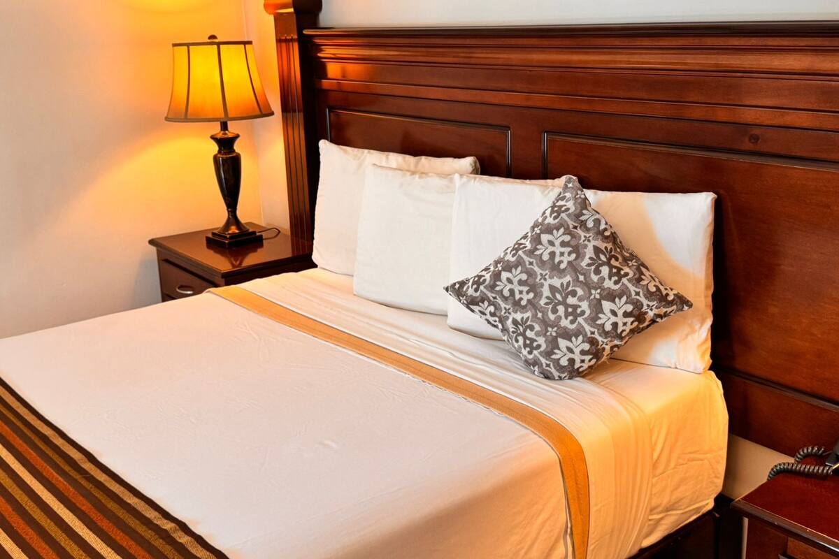 A neatly made bed with white bedding, a patterned pillow, a bedside lamp, and a wooden headboard.