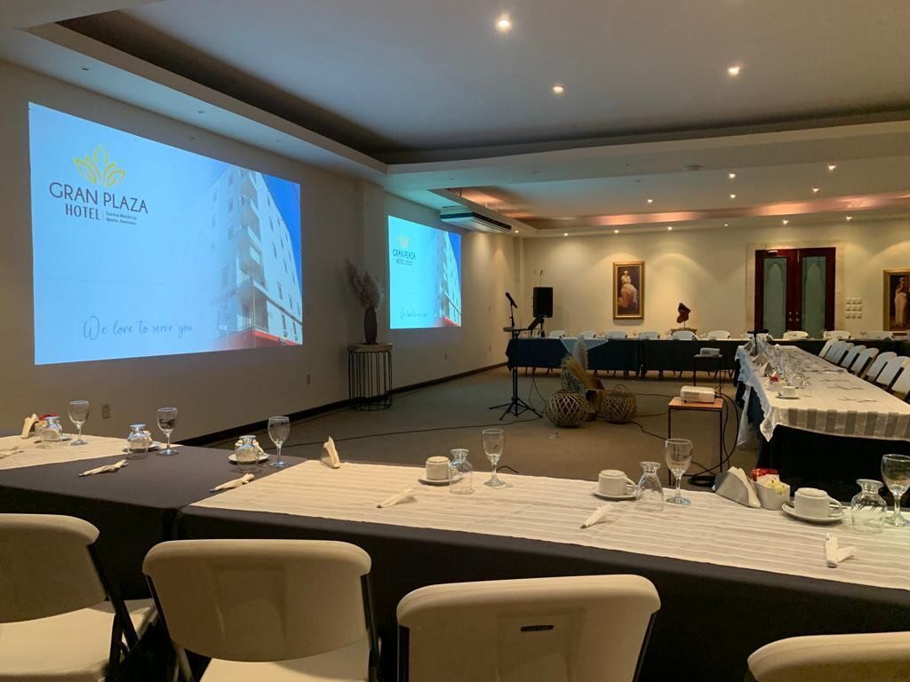 Une salle de conférence avec des tables disposées pour une réunion, un écran de projection affichant le texte « GRAN PLAZA HOTEL », et un système de sonorisation.