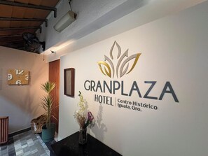 Zona de recepción de un hotel con un reloj, el logotipo del Hotel GranPlaza y un jarrón con flores.