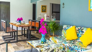 A dining area with a wooden table, chairs, and a vase with pink flowers. There is a sofa with yellow cushions and a patterned throw.
