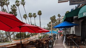 Een buitenontbijtgebied met houten tafels en stoelen, rode en blauwe parasols en palmbomen op de achtergrond.