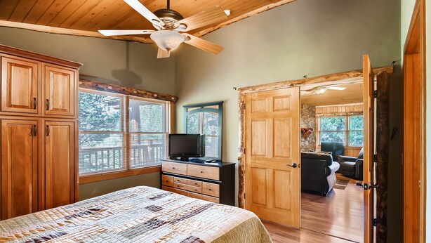 A bedroom with a wooden ceiling fan, a bed with a quilt, a TV on a dresser, and a large wooden door leading to a living area. - Black Canyon Inn Unit A4/5 (Estes Park)