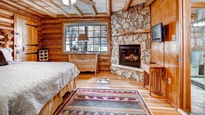 A log cabin bedroom with a stone fireplace, wooden ceiling beams, a bed, a desk, and a television.