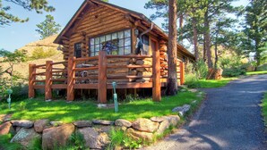 A rustic log cabin with a porch, surrounded by trees and a gravel path.