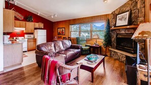 A cozy living room with a fireplace, wooden furniture, and a kitchen area with a red wall and wooden cabinets.