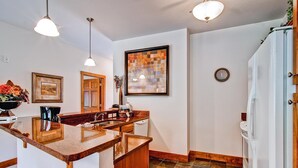 A kitchen with a marble countertop, wooden cabinets, pendant lighting, a clock, and a refrigerator. - Black Canyon Inn Unit F3 (Estes Park)