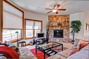 A living room with a stone fireplace, a ceiling fan, and a large window with blinds. - Black Canyon Inn Unit F3 (Estes Park)