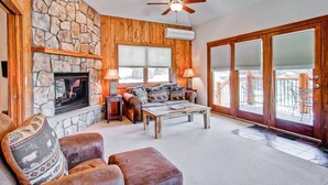 A cozy living room with a stone fireplace, wooden walls, a ceiling fan, and a large sliding glass door leading to an outdoor patio. - Black Canyon Inn Unit F10 (Estes Park)