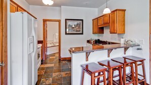A kitchen with wooden cabinets, a white refrigerator, a stone-tiled floor, and a bar area with stools. - Black Canyon Inn Unit F10 (Estes Park)