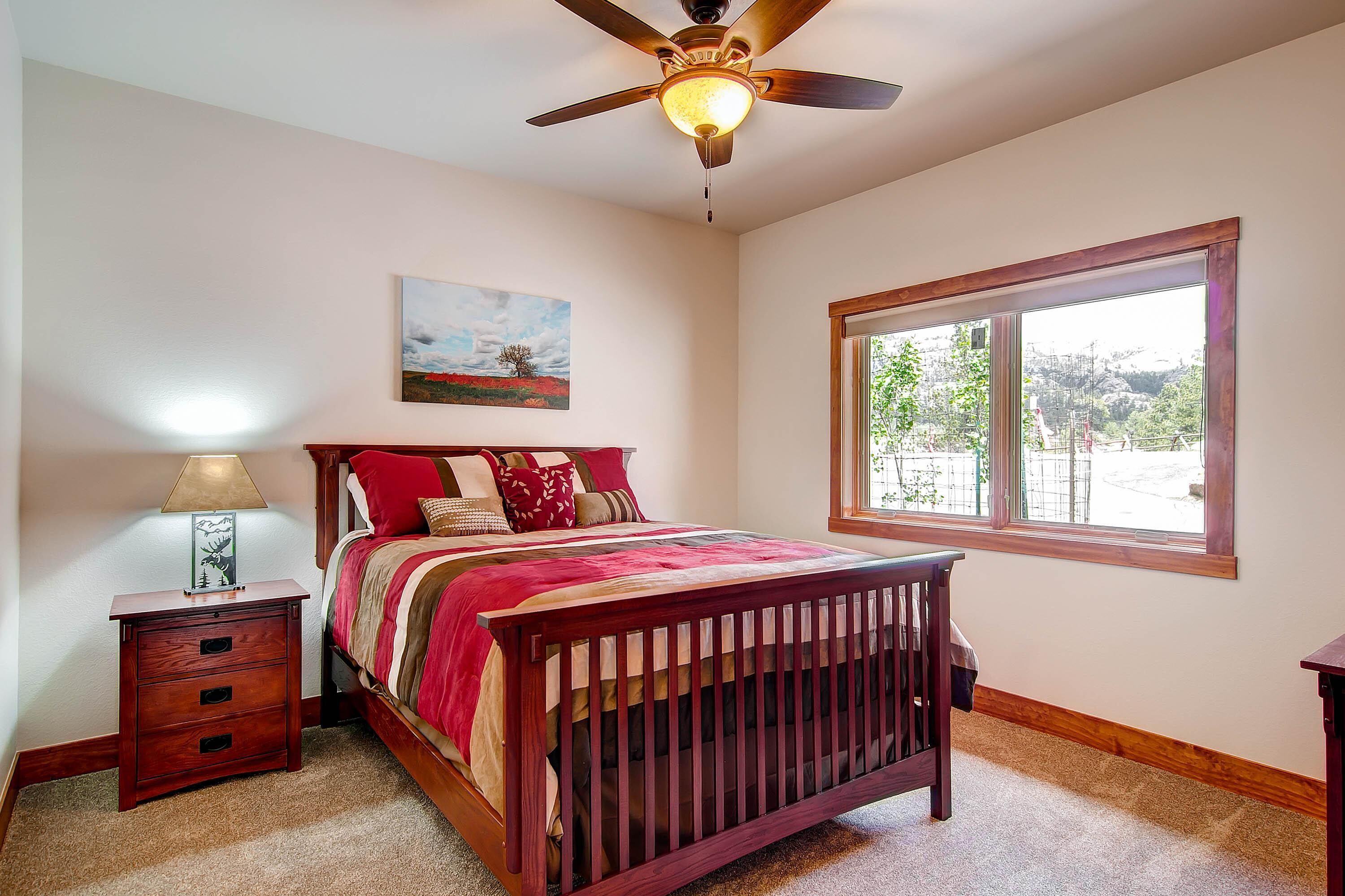A bedroom with a wooden bed, a nightstand, a ceiling fan, a window with a view, and a painting on the wall.
