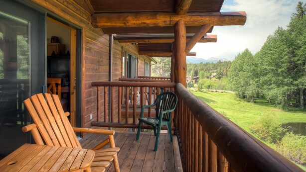 Um deck de madeira com duas cadeiras e uma mesa, com vista para uma paisagem verde exuberante, com árvores e montanhas.
