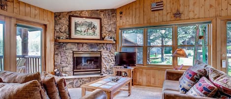 A cozy living room with a stone fireplace, wooden walls, and large windows offering a view of the outdoors.