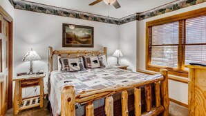 Une chambre à coucher avec un lit en bois, deux tables de chevet avec des lampes, un ventilateur de plafond, un tableau représentant un paysage encadré et une fenêtre avec des stores.