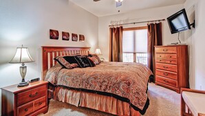 A bedroom with a bed, a nightstand, a dresser, a ceiling fan, a TV mounted on the wall, and a window with curtains. - Black Canyon Inn Unit F7 Deluxe (Estes Park)