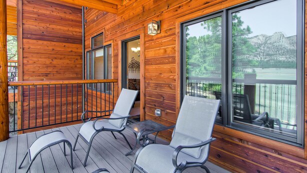 A wooden deck with two chairs and a table, large windows with a view of trees and a lake, and a wooden railing. - Black Canyon Inn Unit F7 Deluxe (Estes Park)