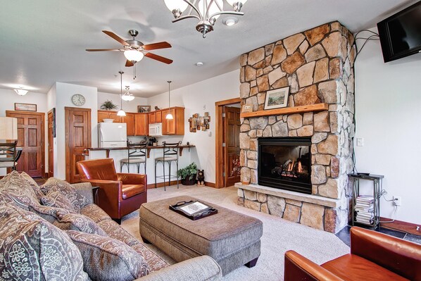 A living room with a stone fireplace, a ceiling fan, and a kitchen in the background.