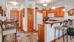A kitchen with wooden cabinets, a refrigerator, a microwave, and a dining area with metal chairs and a table. - Black Canyon Inn Unit F7 Deluxe (Estes Park)
