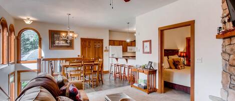 A spacious living room with a ceiling fan, a dining area with wooden furniture, a kitchen with a stone fireplace, and a bedroom with a bed and a nightstand.