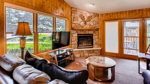 A cozy living room with wooden walls, a stone fireplace, a flat-screen TV, leather sofas, and a large window offering a view of the outdoors. - Black Canyon Inn Unit B4/5 (Estes Park)