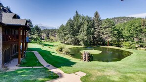 A lodge with a stone exterior and wooden porch, a well-maintained lawn, a pond, and a mountain backdrop. - Black Canyon Inn Unit B4/5 (Estes Park)
