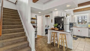 Eine Küche mit Kochinsel, Holzbarhocker, einem Kühlschrank und einem Fenster mit Jalousien.
