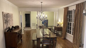 A dining area with a long wooden table, chairs, a chandelier, and a view of a room with a window and curtains.