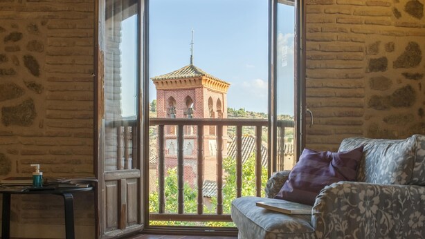 A room with a wooden floor, a patterned sofa, a small table with a bottle, and a view of a brick tower through a glass door. - San Cipriano. Apartment with garage included (Toledo)