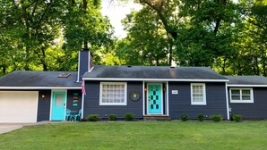 Une maison à deux étages, à l’extérieur sombre, avec une porte d’entrée turquoise et un petit jardin devant, composé d’une pelouse et de quelques arbustes.