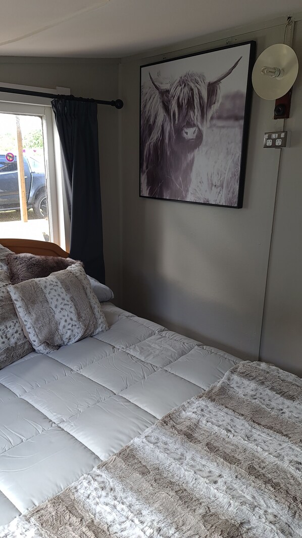 A bedroom with a bed, a window with curtains, a wall-mounted light fixture, and a painting of a yak. - Cute rustic bach, A home away from home. Located in beautiful mangakino (Waikato)