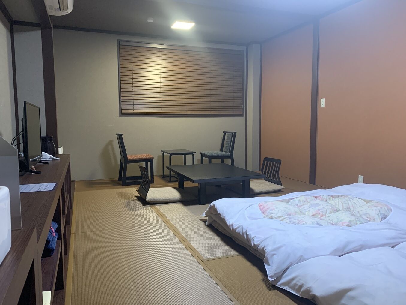 A room with a bed, a desk with a computer, two chairs, a table, and a window with blinds.