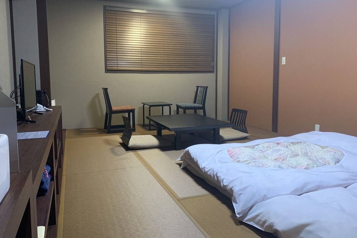 A room with a bed, a desk with a computer, two chairs, a table, and a window with blinds.