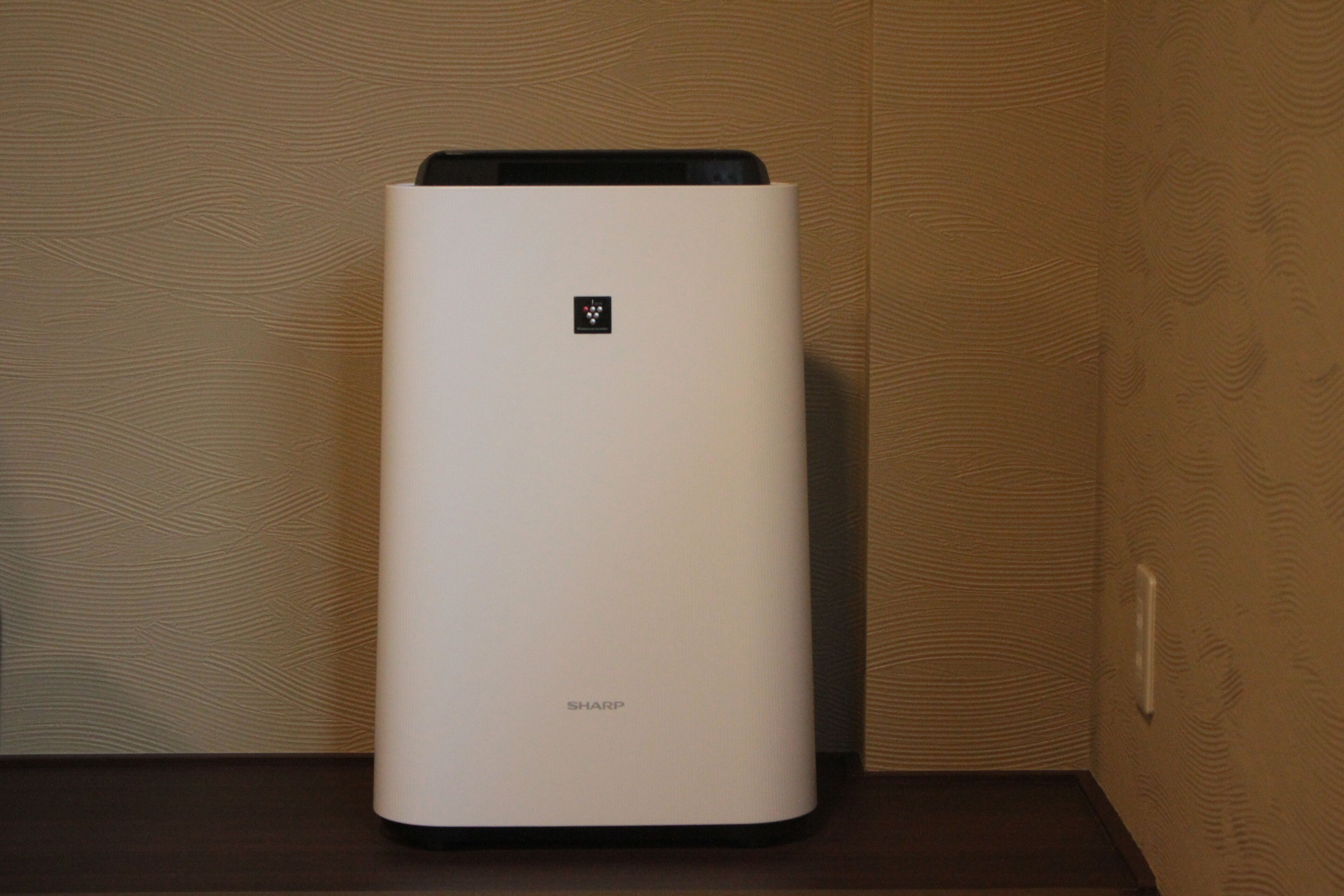 A white, modern air purifier with the brand name 'SHARP' on a wooden surface against a wooden wall.