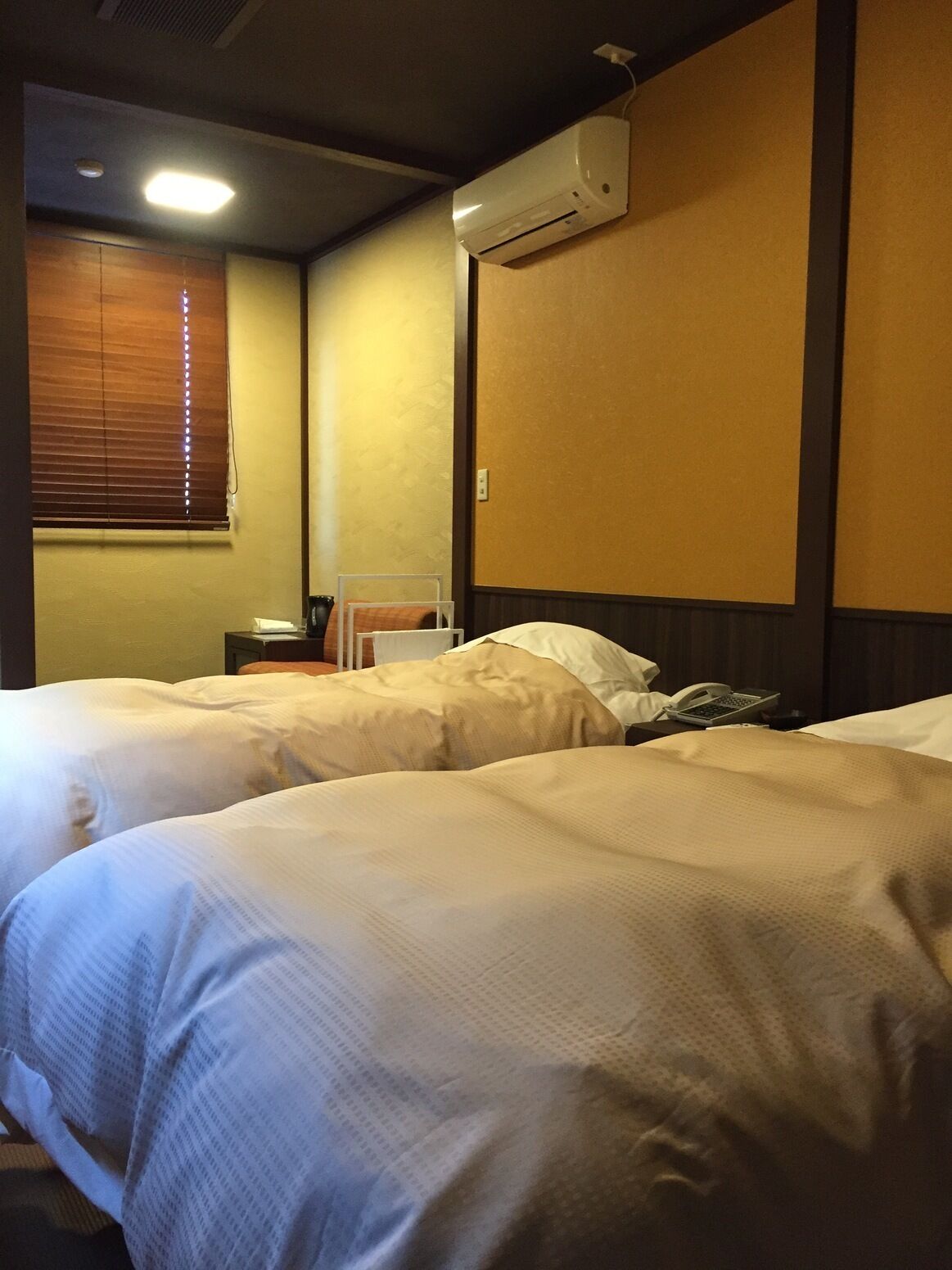 A hotel room with two beds, an air conditioning unit, a bedside table with a phone, and a window with blinds.