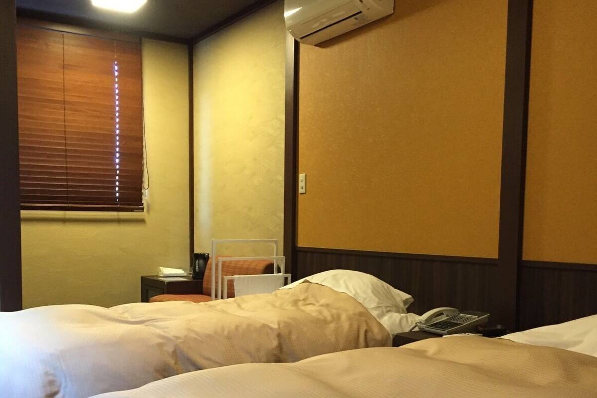 A hotel room with two beds, an air conditioning unit, a bedside table with a phone, and a window with blinds.