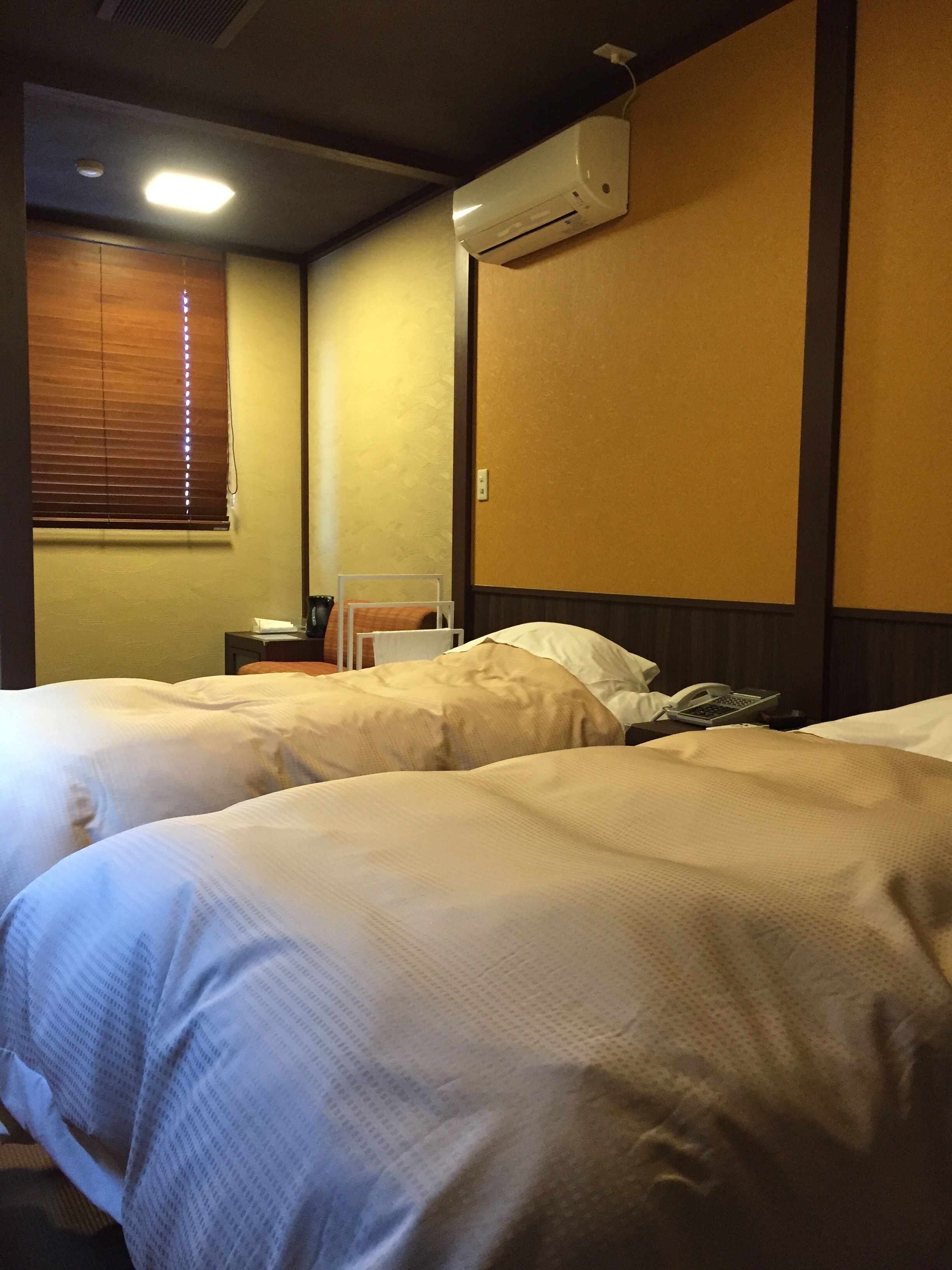 A hotel room with two beds, a bedside table with a phone and a lamp, an air conditioning unit on the wall, and a window with blinds.