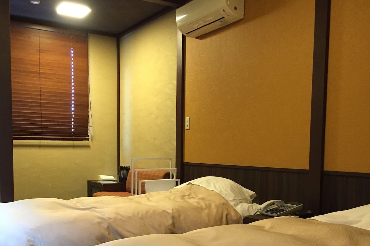 A hotel room with two beds, a bedside table with a phone and a lamp, an air conditioning unit on the wall, and a window with blinds.