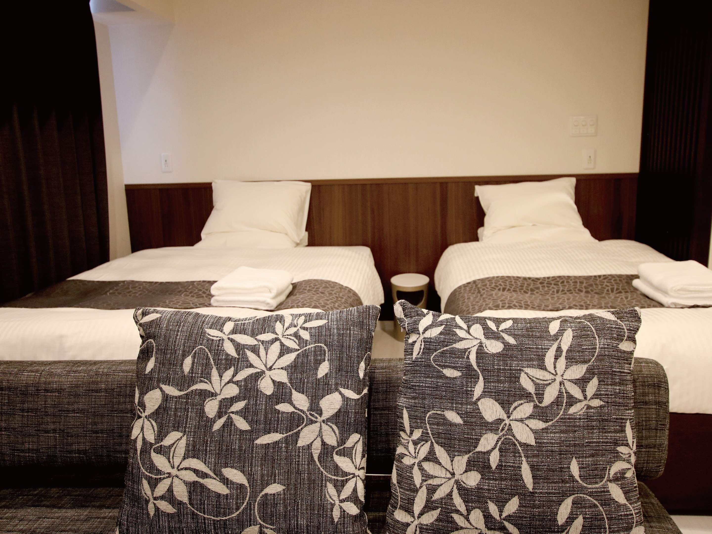 Una habitación de hotel con dos camas, cada una con ropa de cama blanca y cabeceros marrones. Hay dos mesitas de noche con lámparas, y la habitación tiene un cabecero de madera.