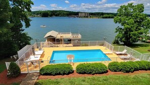 Une piscine avec vue sur un lac, entourée d’une terrasse délimitée par une clôture, comprenant des chaises longues et un petit bâtiment.
