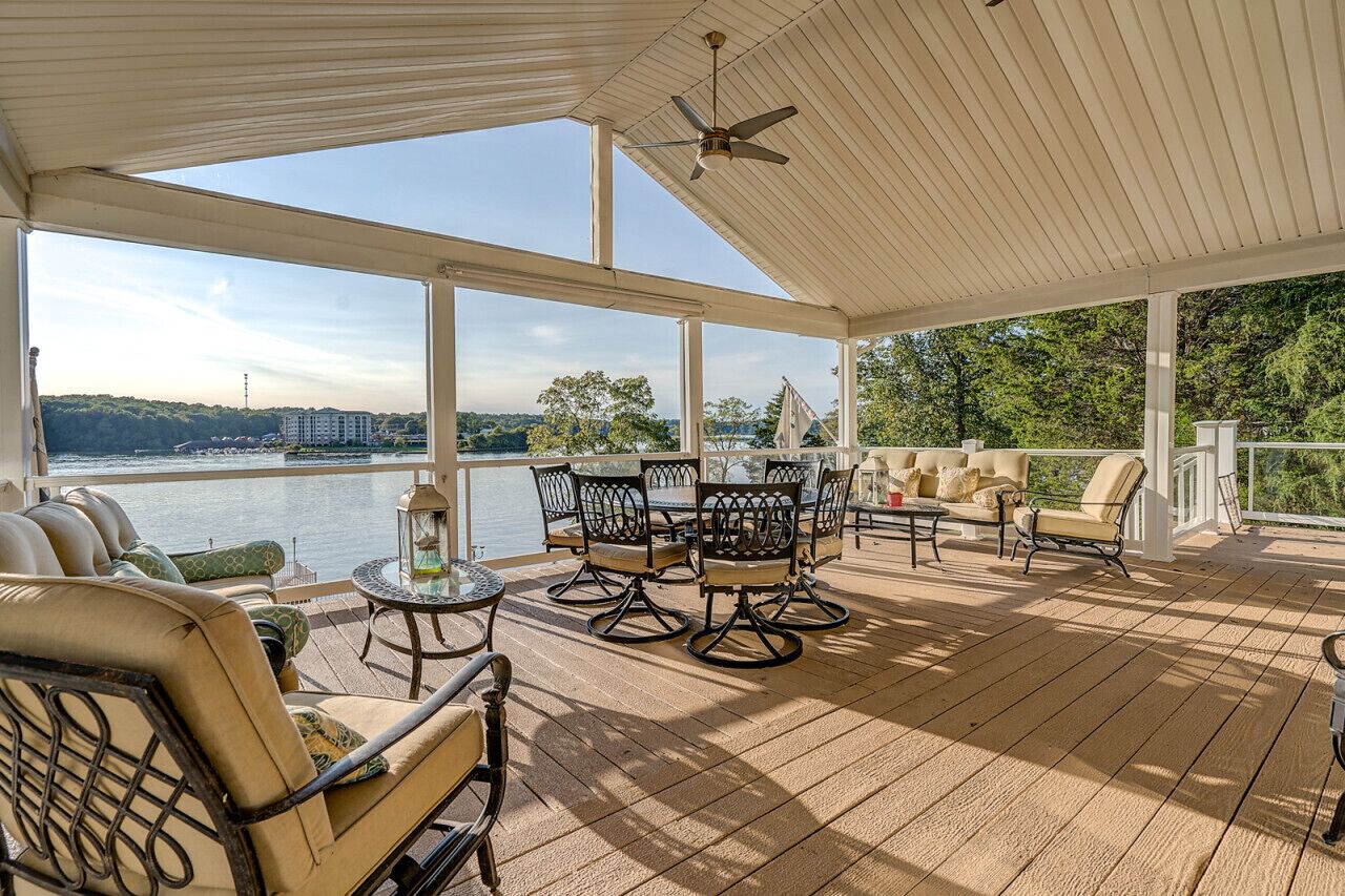 A covered outdoor deck with seating and tables, overlooking a body of water and greenery.
