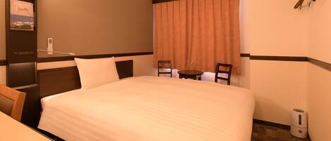 A hotel room with a large bed, a desk, a chair, a small table, a window with curtains, a wall-mounted telephone, and a trash bin.