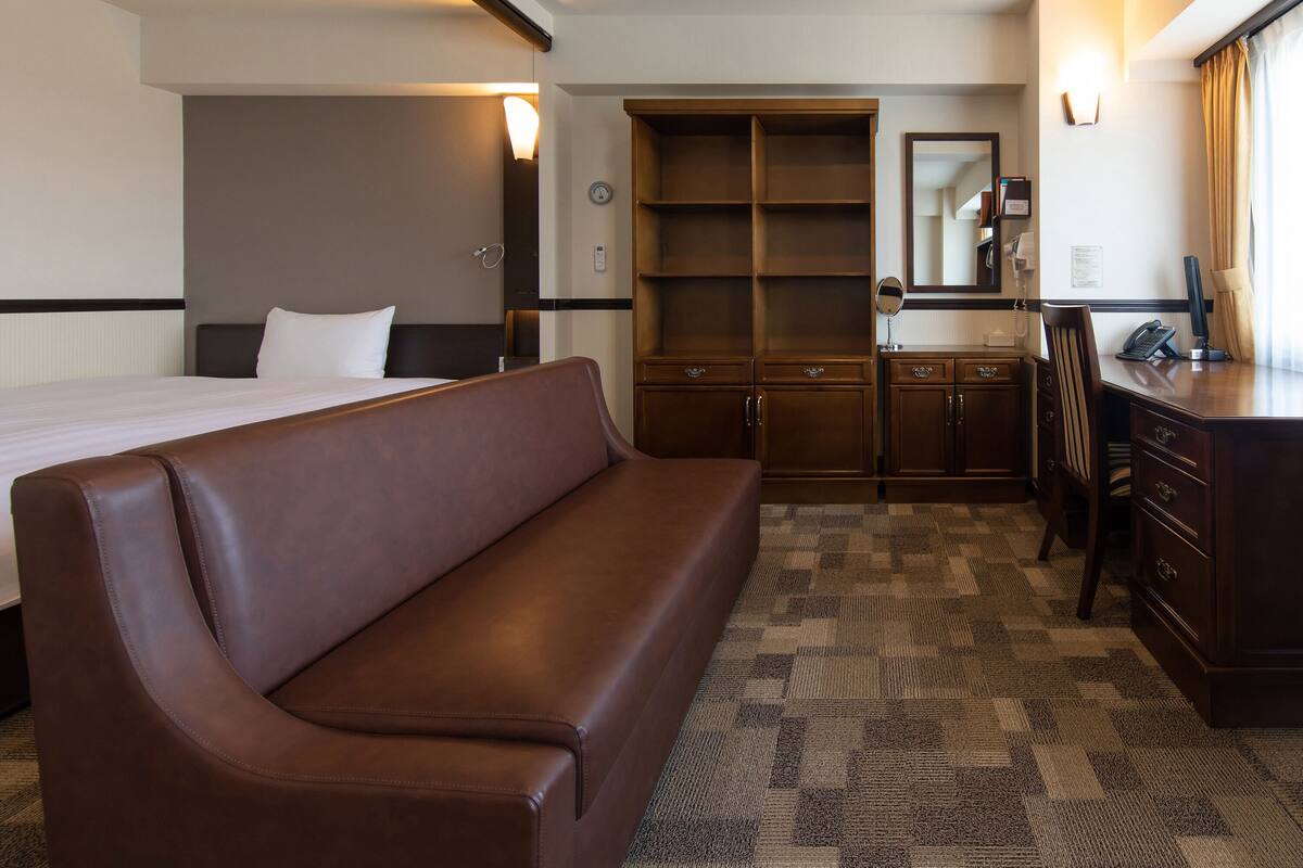 A hotel room with a bed, a brown sofa, a desk with a telephone, a mirror, and a wardrobe.
