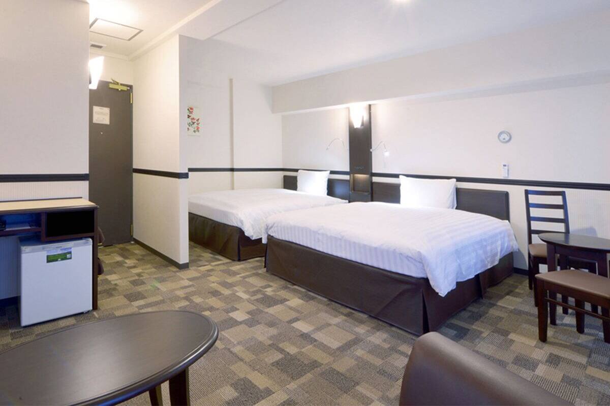 A hotel room with two single beds, a desk, a chair, a small table, and a refrigerator.