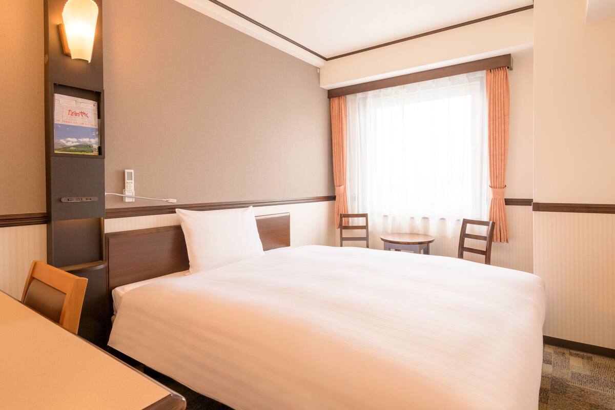 A hotel room with a large bed, a desk, a chair, a small table, and a window with curtains.