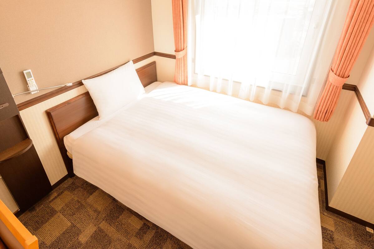 A neatly made bed in a hotel room with a wooden headboard, a bedside table with a lamp, and a window with sheer curtains.