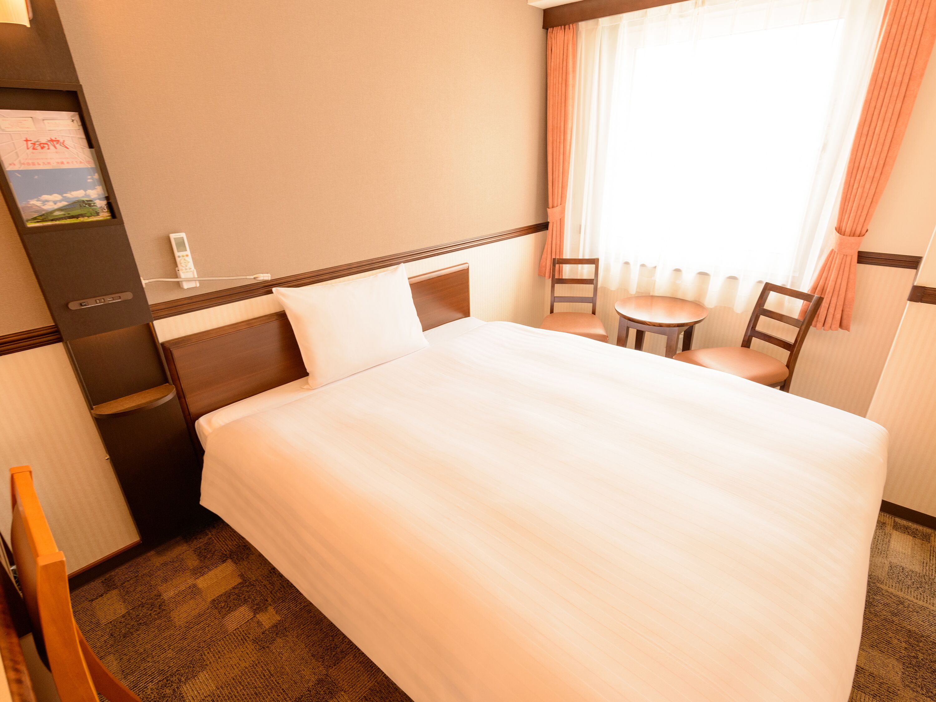 Ein Hotelzimmer mit einem großen Bett, einem kleinen Tisch, zwei Stühlen, einem Schreibtisch mit Telefon und einem Fenster mit Vorhängen.