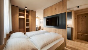 A modern bedroom with two beds, wooden paneling, a flat-screen TV, and a small kitchenette. - Apartments Alpe di Susi - Holiday Apartments in the Italian Dolomites (Castelrotto)