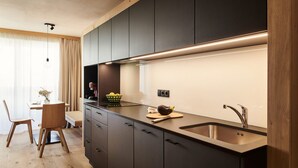 A modern kitchen with dark cabinetry, a stainless steel sink, and a wooden dining area with a table and chairs. - Apartments Alpe di Susi - Holiday Apartments in the Italian Dolomites (Castelrotto)