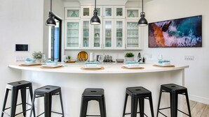 Una cocina moderna con armarios blancos, taburetes negros y una isla grande con cristalería y elementos decorativos.