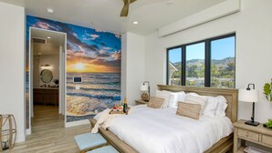 Un dormitorio con una cama grande, vista al océano por una ventana, un ventilador de techo y un mural de un atardecer.