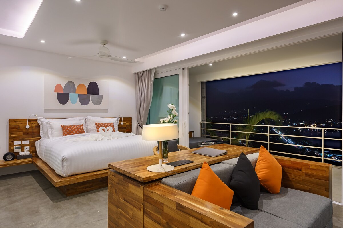 A modern hotel room with a large bed, a sofa, a desk, and a view of the city at night.